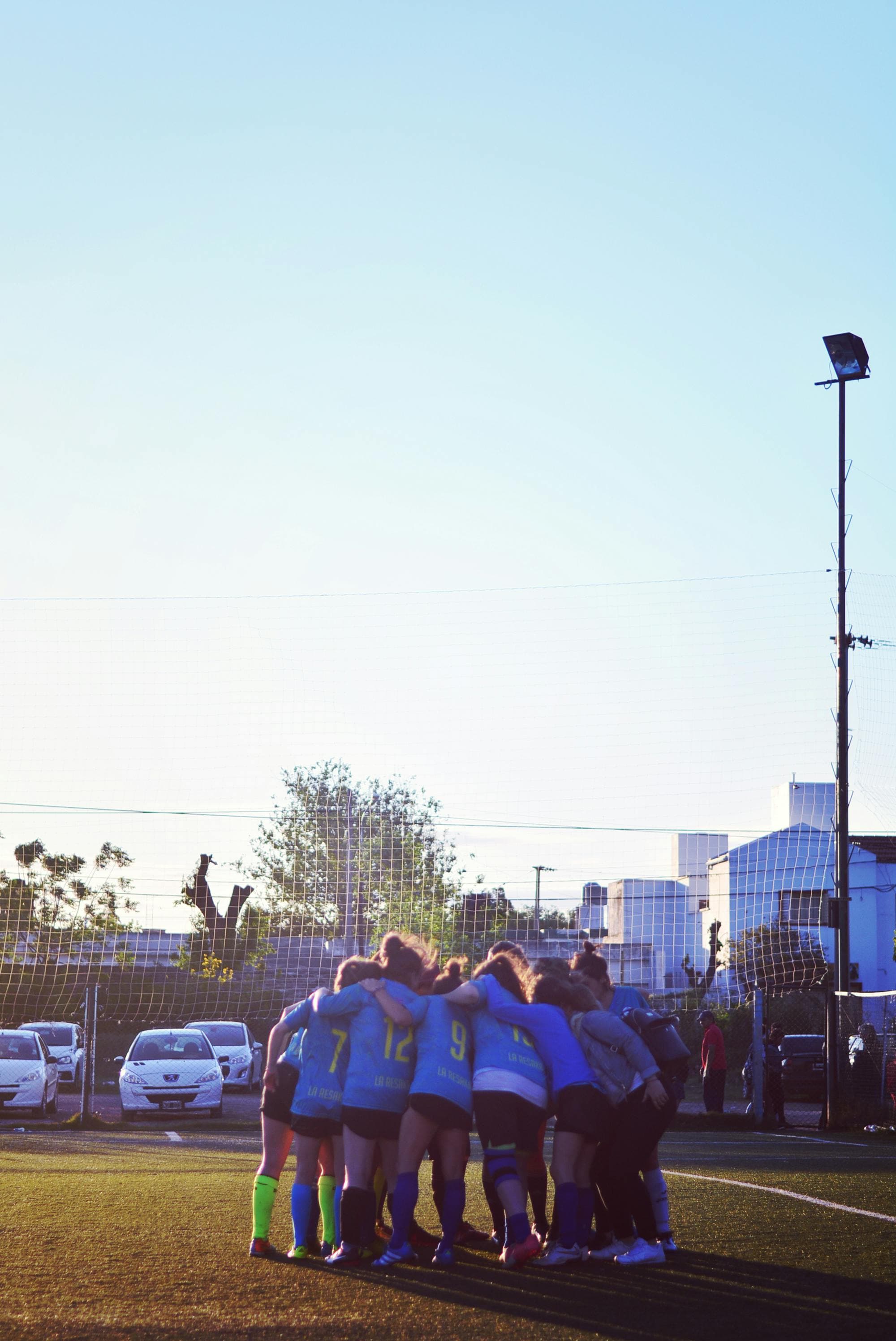 An adult soccer team gathered in a pre-match huddle on an outdoor field.